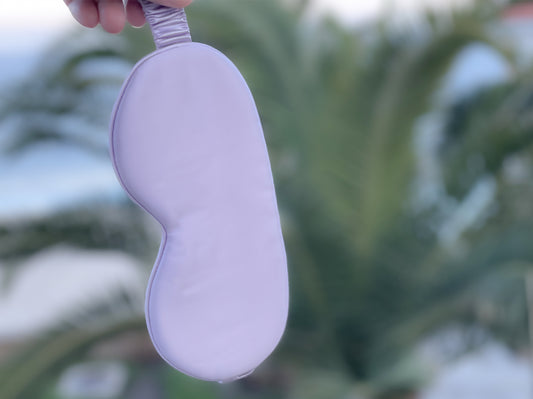 Lilac silk eye mask for sleeping held in front of palm trees - made from organic silk